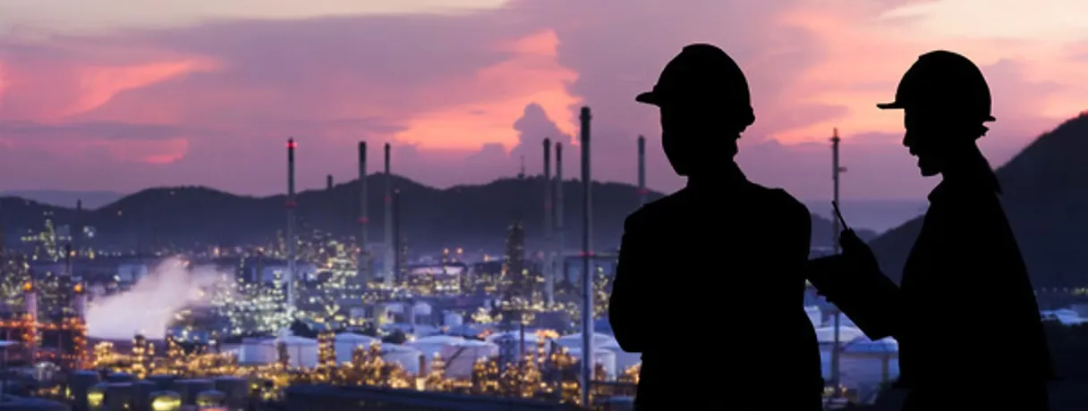 Silhouettes of two people in hard hats overlooking a lit refinery and hills under a pink dusk sky.